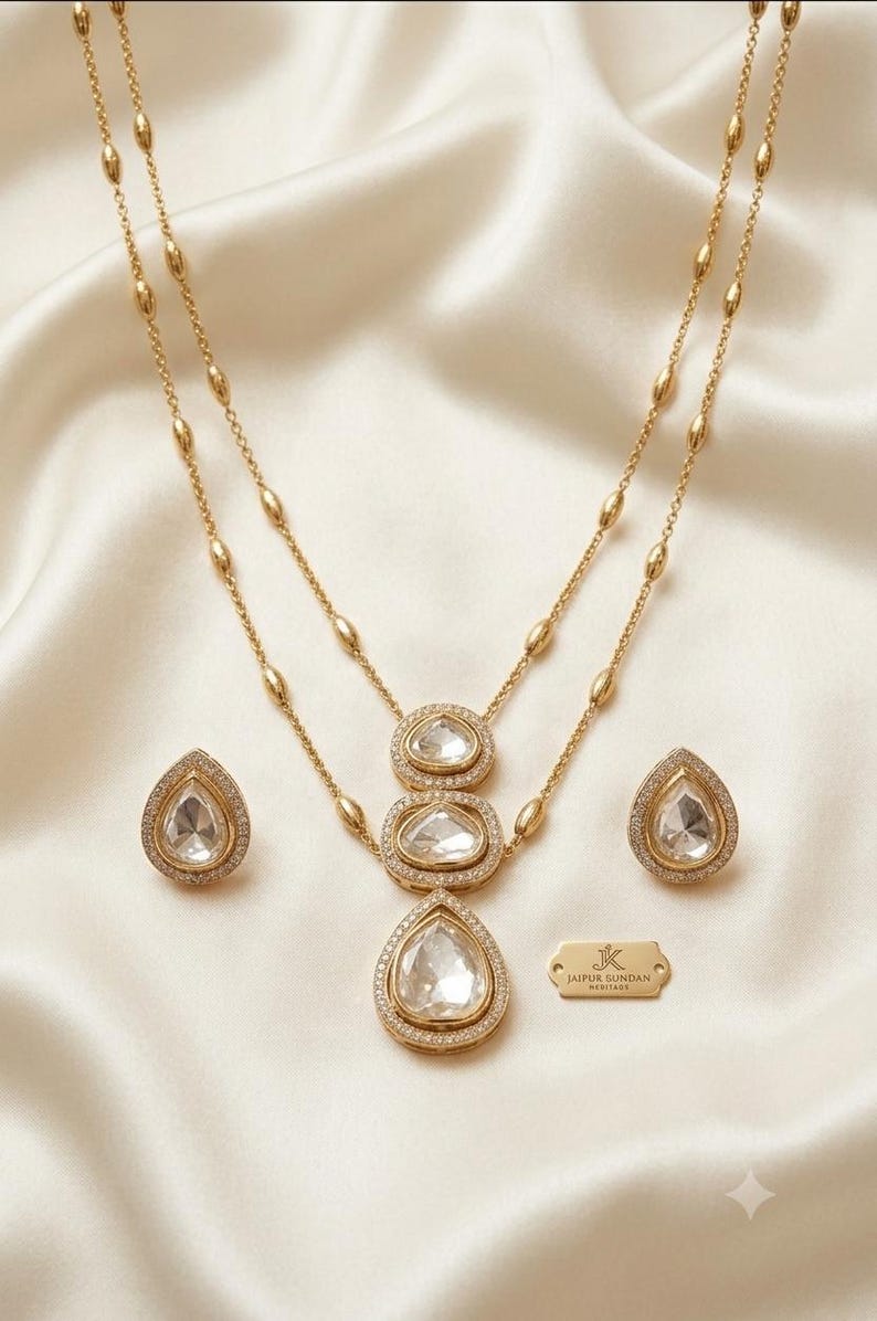 May include: A gold-toned necklace and earring set featuring clear, pear-shaped stones surrounded by small white crystals. The necklace has a double-layered chain with oval accents and a pendant with three stacked stones. A small gold tag reads JK JAIPUR SUNDAN HERITAGE.