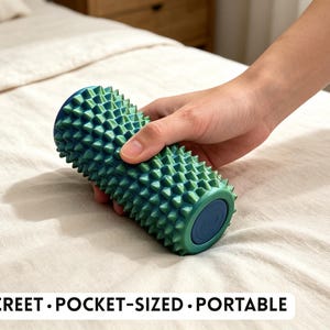 Könnte beinhalten: Eine zylindrische Massagerolle mit einer strukturierten Oberfläche aus zahlreichen kleinen, spitzen Noppen. Die Rolle ist in einem Farbverlauf von Petrol und Grün gehalten und hat eine dunkelblaue Kappe. Der Text lautet: DISCREET • POCKET-SIZED • PORTABLE.