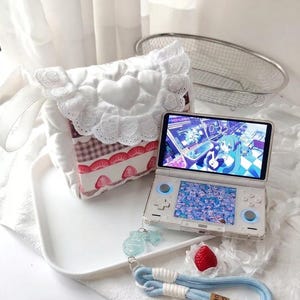 May include: A white quilted bag with a heart-shaped flap and lace trim, paired with a handheld gaming console featuring a blue-haired anime character on the screen, a blue wrist strap with a wooden tag, and a small red strawberry decoration.