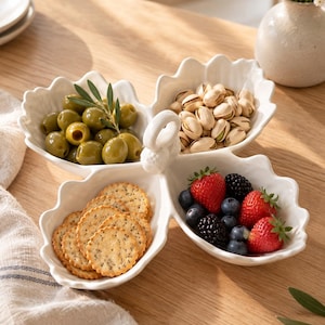 May include: A white ceramic three-section serving dish shaped like leaves with a central loop handle. The sections are filled with green olives, shelled pistachios, round crackers with poppy seeds, and fresh strawberries and blackberries.