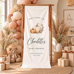 May include: A vertical cream-colored fabric banner hanging on a wooden stand, featuring the text Welcome to Charlotte's Baby Shower, Baby Charlotte, and 10.17.2026, decorated with a watercolor illustration of two pumpkins and autumn foliage.