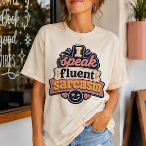 Könnte beinhalten: Ein cremefarbenes Kurzarm-T-Shirt mit einer mehrschichtigen Stickerei auf der Brust. Das Design zeigt den Text "I speak fluent sarcasm" in gestapelten, stilisierten Buchstaben sowie ein zwinkerndes Smiley-Gesicht am unteren Rand.