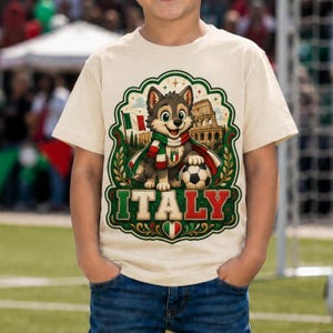 May include: A cream-coloured t-shirt featuring a graphic of a cartoon wolf wearing a scarf with the colours of the Italian flag. The wolf stands next to a football and the Colosseum. The word ITALY is printed in large letters below the design.