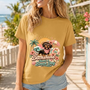 May include: A mustard yellow t-shirt featuring a graphic of a dachshund wearing heart-shaped sunglasses and a pink bandana. The design includes a palm tree, beach umbrella, ocean waves, and the text My Dachshund Needs a Beach Day.