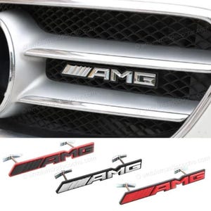 Puede incluir: Tres emblemas de coche AMG con herrajes de montaje de metal. Un emblema es rojo con letras negras, otro es plateado con letras negras y otro es rojo con letras negras. Los emblemas muestran las letras AMG y un gráfico de rayas.