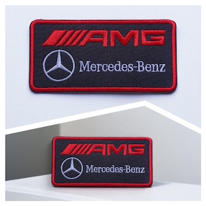 Puede incluir: Un parche rectangular bordado con fondo negro y borde rojo. El diseño incluye el logotipo rojo de AMG, el emblema plateado de la estrella de tres puntas de Mercedes-Benz y el texto Mercedes-Benz bordado en blanco sobre la tela.