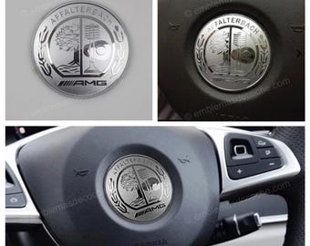 Puede incluir: Un emblema metálico circular de color plateado que presenta el escudo de Affalterbach con un diseño de árbol y un resorte de válvula, coronado con el texto AFFALTERBACH y el logotipo de AMG en la parte inferior, para el volante del vehículo.
