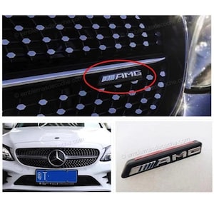 Puede incluir: Un emblema AMG negro y plateado diseñado para la rejilla de un vehículo. La insignia rectangular presenta las letras AMG en un acabado metálico sobre un fondo negro, mostrada tanto instalada en la rejilla de un coche como pieza suelta.