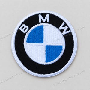 Puede incluir: Un parche bordado circular que presenta el logotipo de BMW. El diseño incluye un anillo exterior negro con las letras blancas BMW, rodeando un círculo central dividido en cuatro cuadrantes de colores azul y blanco alternos.