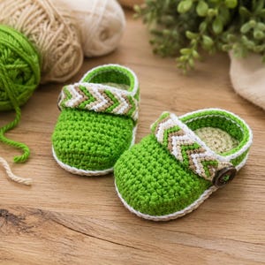 May include: A pair of handmade crochet baby shoes in a vibrant green color. Each shoe features a white and tan chevron patterned strap across the top, secured with a dark brown circular button on the side. The edges are finished with white trim.