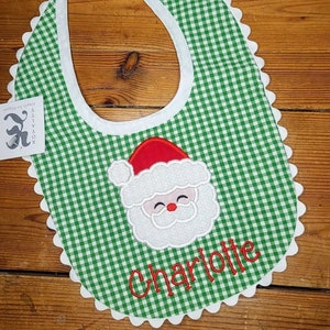 May include: Green and white gingham baby bib with a white scalloped edge. The bib features an embroidered Santa Claus face and the name "Charlotte".