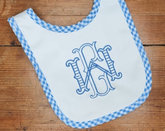 May include: A white baby bib with a blue gingham trim and a blue embroidered monogram. The monogram is a traditional script style with the letters "W G" intertwined.