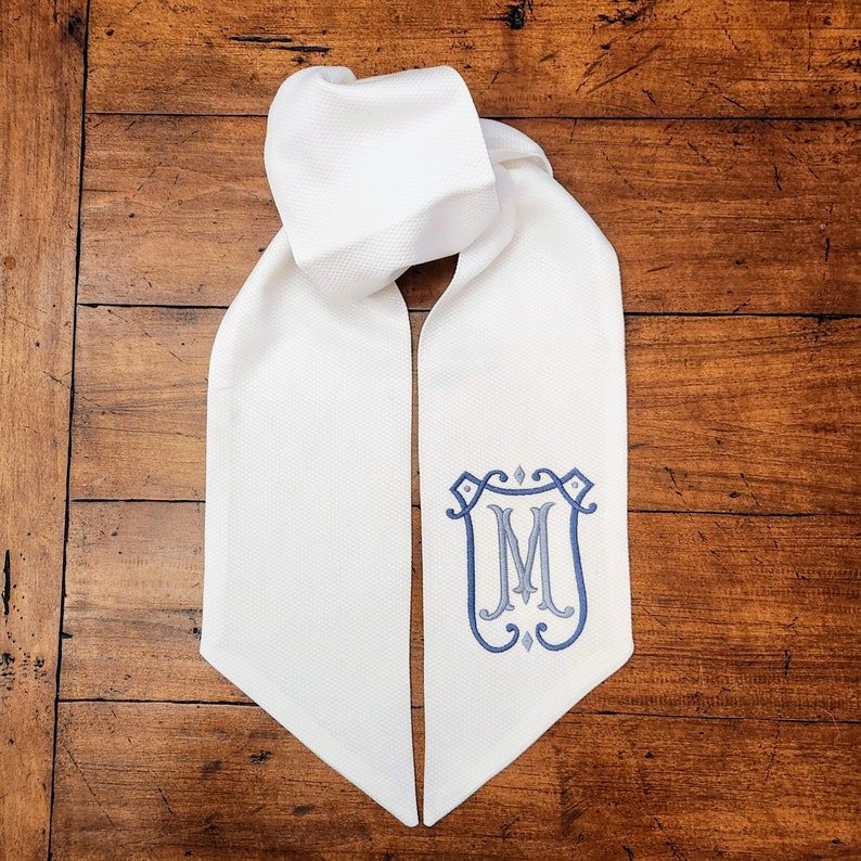 May include: White graduation stole with a blue and gray embroidered monogram of the letter "M" in a decorative frame.