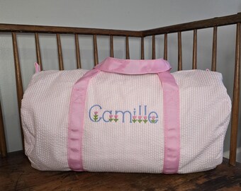 May include: A pink and white gingham duffel bag with pink straps and the name "Camille" embroidered in blue with pink flowers.