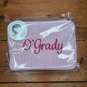 May include: A pink and white striped fabric pouch with the name "O'Grady" embroidered in pink. The pouch is packaged in clear plastic.