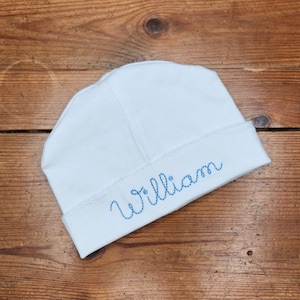 May include: White baby hat with blue embroidered lettering that says "William".