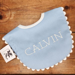 May include: A light blue baby bib with a white scalloped edge and the name "CALVIN" embroidered in white.