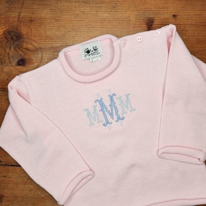 May include: A pink knitted baby sweater with a blue embroidered monogram "MMM" on the front.