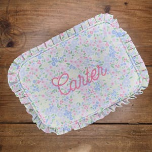 May include: A rectangular, floral-patterned baby pillow with a ruffled edge. The pillow features a light blue, pink, and yellow floral design. The name "Carter" is embroidered in pink cursive across the center.