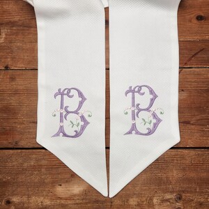 May include: Two white fabric table runners with a purple embroidered letter "B" with floral accents on each runner.