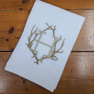 May include: White kitchen towel with a brown embroidered monogram of the letter H inside a wreath of antlers.