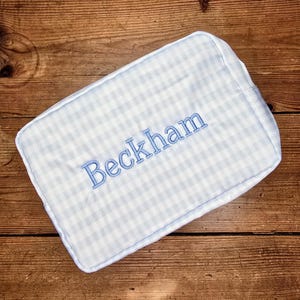 May include: A rectangular, light blue and white gingham toiletry bag with the name "Beckham" embroidered in light blue thread. The bag is set against a wooden background.