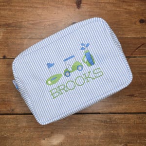 May include: A seersucker golf-themed travel bag with blue and white stripes. The bag features embroidered golf course elements, including a golf cart, flag, and golf bag. The name "BROOKS" is embroidered in green.