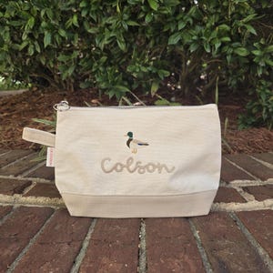 May include: A beige canvas zippered pouch with a brown duck and the name "Colson" embroidered on it.