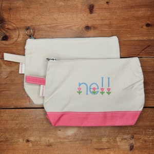 May include: Two canvas zippered pouches, one nested inside the other. The smaller pouch has a pink bottom and the words "nell" embroidered in blue with pink tulips. Both pouches have a red and white label that says "CB Station".