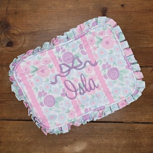 May include: Rectangular floral print pillow with the name "Isla" embroidered in purple script. The pillow features a ruffled edge and a pattern of pink stripes and flowers in shades of pink, purple, and blue.