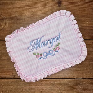 May include: Pink and white gingham baby blanket with the name "Margot" embroidered in light blue script. The blanket has a ruffled edge and a floral and bow design.