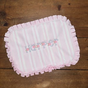 May include: A rectangular, pink and white striped baby blanket with a ruffled edge. The name "Harper" is embroidered in light blue with pink flower accents. The blanket is made of a soft, likely cotton material.