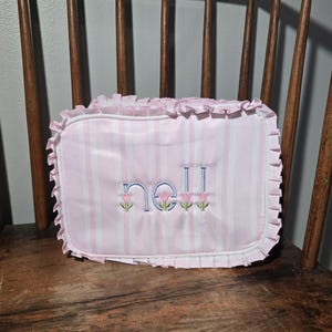 May include: A rectangular pink and white striped pillow with the name "Nell" embroidered in blue thread. The pillow has a ruffled edge and is decorated with small pink flower accents.
