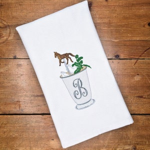 May include: White kitchen towel with an embroidered design of a cocktail glass with a horse on a straw, ice cubes, a green leaf, and the letter "B" in a gray font.