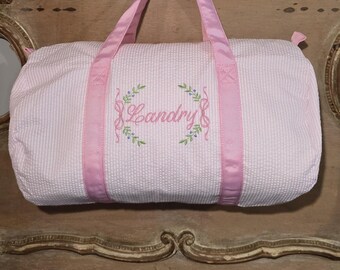May include: A pink seersucker duffel bag with pink satin handles and trim. The bag features the embroidered name "Landry" in pink script, surrounded by a floral wreath design. The bag is designed for carrying items.