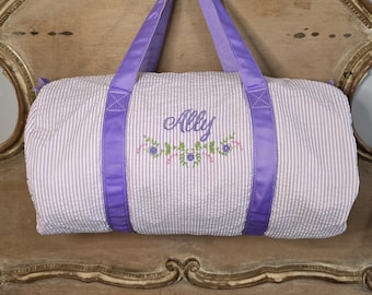 May include: A seersucker duffel bag with vertical stripes in white and light purple. The bag has purple handles and trim. The name "Ally" is embroidered in cursive with floral accents.