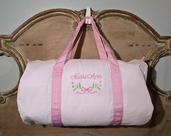 May include: A white seersucker duffel bag with pink accents. The bag features pink handles and trim, with the name "Norri Ann" embroidered in pink script, surrounded by a floral design. The bag is hanging on a white hook.