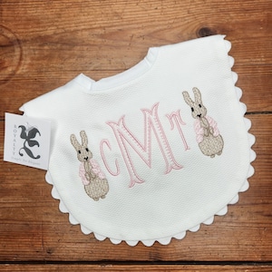 May include: White baby bib with scalloped edges, featuring two embroidered brown bunnies wearing pink jackets and the monogram "CMT" in pink thread.
