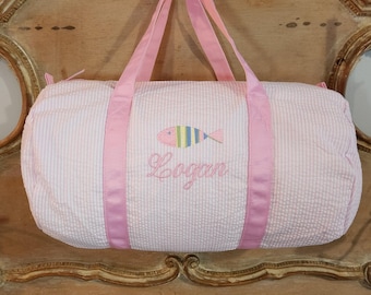 May include: Pink and white striped duffel bag with pink straps and a personalized embroidered fish and the name "Logan".