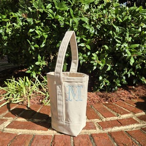 May include: A white canvas tote bag with a blue embroidered letter "M" on the front. The bag has a single handle and is sitting on a brick surface.