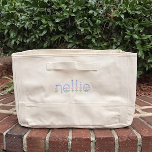 May include: A rectangular off-white canvas storage bin featuring a fabric handle on the front. The name "nellie" is embroidered in blue lowercase letters, with small pink and green tulip-like floral motifs positioned beneath each letter.