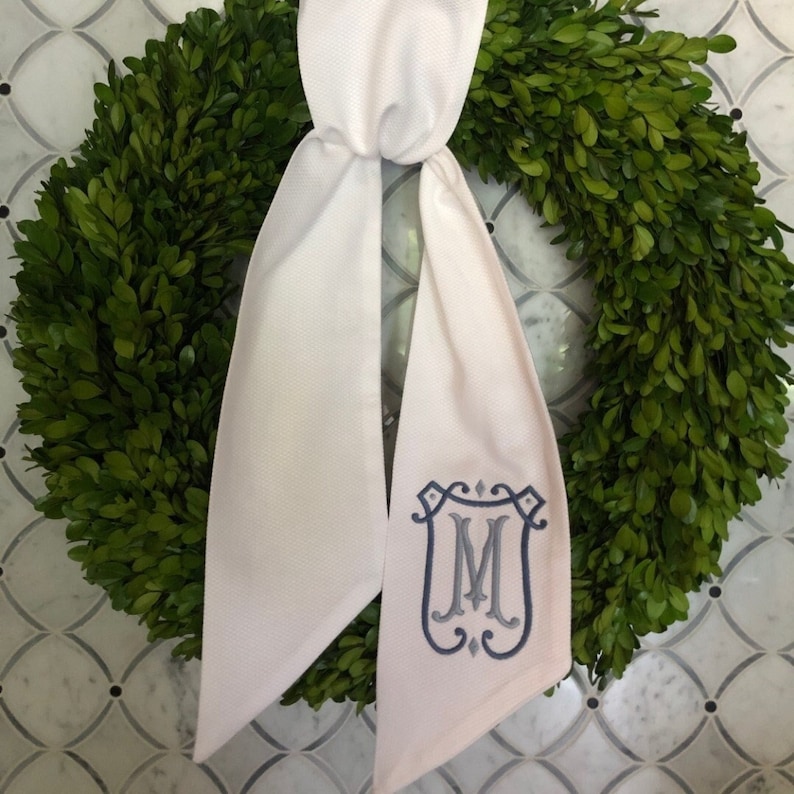 May include: A white fabric bow with a blue embroidered monogram "M" in a decorative frame. The bow is tied around a green wreath.