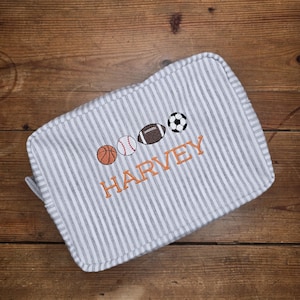 May include: A gray and white striped travel bag with embroidered sports balls and the name "HARVEY" in orange thread. The bag features a basketball, baseball, football, and soccer ball design.