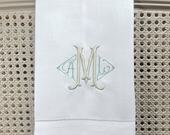 May include: White linen hand towel with a light blue and cream embroidered monogram "AML" in a diamond shape.