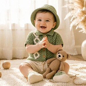 May include: A baby wearing a sage green crocheted bucket hat, a matching short-sleeved collared shirt with a white diamond pattern, and cream-colored knit shorts and booties. The baby is holding a small brown crocheted teddy bear toy.