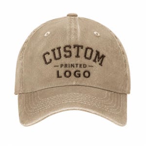 May include: A tan baseball cap with a distressed, washed-out finish. The front features dark brown embroidered text that reads CUSTOM PRINTED LOGO in a bold, arched font style with a horizontal line separating the two words.