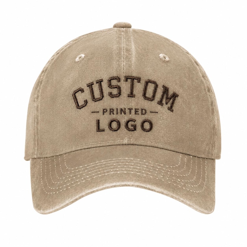 May include: A tan baseball cap with a distressed, washed-out finish. The front features dark brown embroidered text that reads CUSTOM PRINTED LOGO in a bold, arched font style with a horizontal line separating the two words.