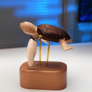 May include: A wooden sea turtle figurine featuring a light-toned head and flippers with a dark brown shell. The turtle is mounted on two thin brass rods attached to a smooth, rounded rectangular wooden base, creating a suspended effect.