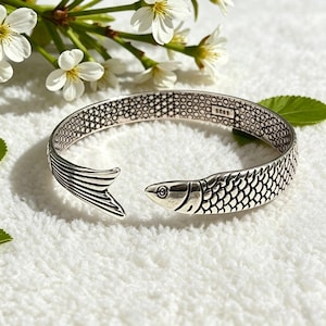 May include: A silver cuff bracelet shaped like a fish with a textured scale pattern on the exterior and a honeycomb pattern on the interior. The bracelet features a fish head at one end and a tail at the other, with the hallmark S925 stamped inside.