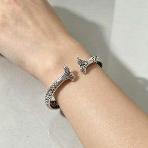 May include: A silver cuff bracelet featuring a textured fish scale pattern along the band and two flared, fin-shaped terminals at the open ends. The metal has an oxidized finish that highlights the intricate details of the scales and the curved edges.